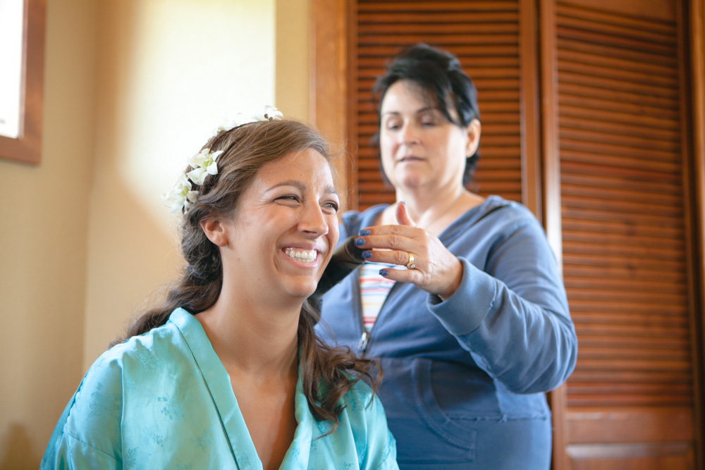 A woman in a blue robe smiles as another woman styles her hair, preparing her for a special occasion. The scene is indoors, with wooden shutters in the background. Jessica Hill Photography - Wedding Photographer - Oregon