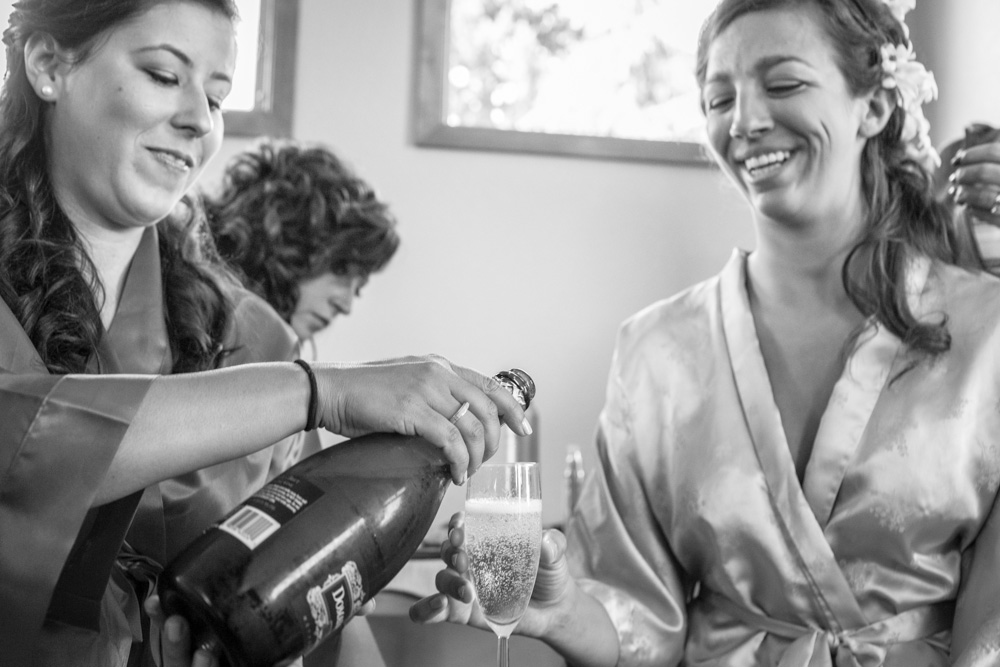 Two women in satin robes smile as one pours champagne into a glass. Another woman sits in the background. The scene appears joyful and celebratory, likely part of a special occasion. Jessica Hill Photography - Wedding Photographer - Oregon
