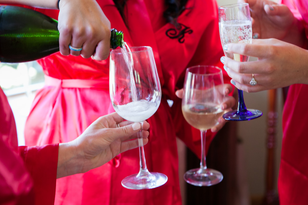 Three people in matching red robes hold wine glasses as one pours sparkling wine into a glass. The scene suggests a celebratory moment, possibly before a special event. Jessica Hill Photography - Wedding Photographer - Oregon