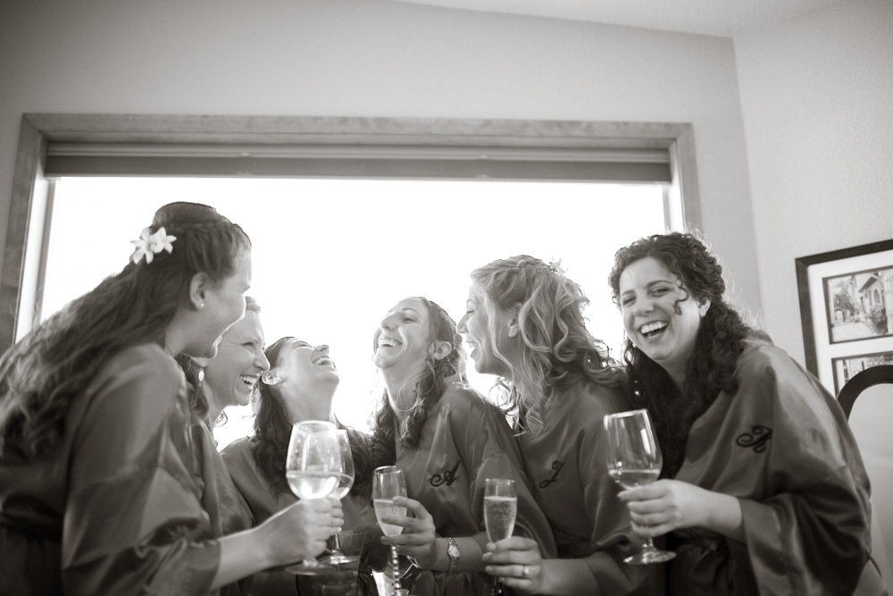 Six women in matching robes stand together indoors, laughing and holding wine glasses. Sunlight streams through a window behind them, creating a joyful and celebratory atmosphere. Jessica Hill Photography - Wedding Photographer - Oregon