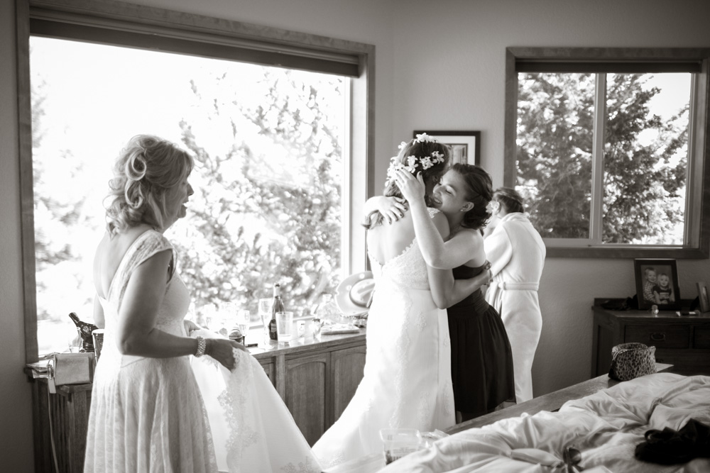 A bride in a wedding dress hugs a bridesmaid in a warmly lit room, while another woman holds the bride's train. A fourth woman stands by the window. The room appears cozy and filled with natural light. Jessica Hill Photography - Wedding Photographer - Oregon