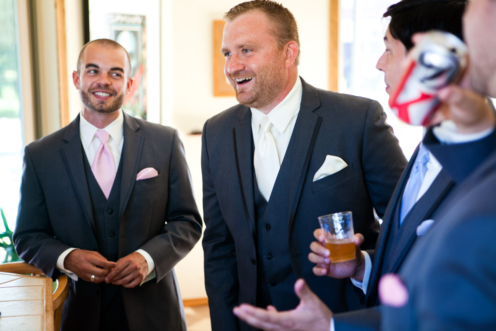Three men in suits and ties are standing indoors, smiling and talking. One holds a beer glass, another has a can. They seem relaxed and happy, likely at a formal event or wedding. Jessica Hill Photography - Wedding Photographer - Oregon