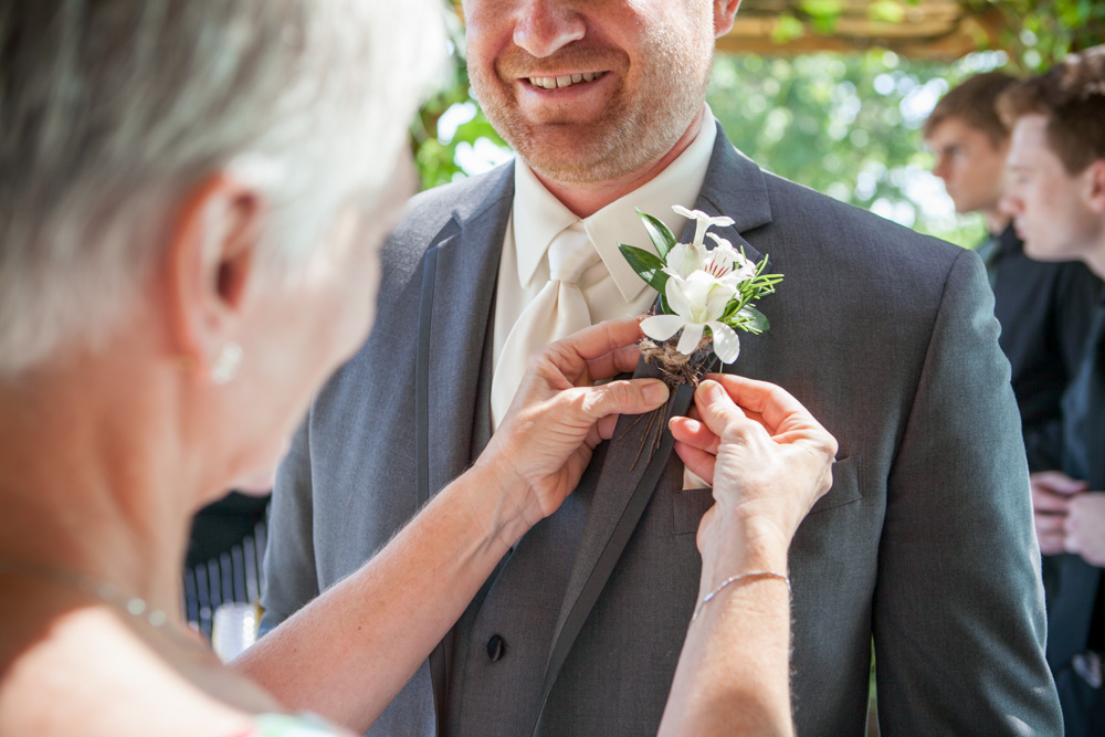 A woman pins a white flower boutonniere on a smiling man's suit jacket at an outdoor event, with other people blurred in the background. Jessica Hill Photography - Wedding Photographer - Oregon