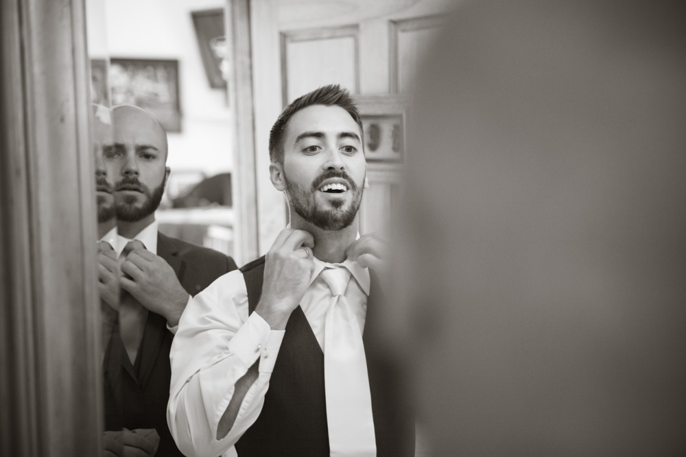 A man in a suit adjusts his tie in front of a mirror, smiling, with another man behind him also fixing his tie. The image is black and white, capturing a candid preparation moment. Jessica Hill Photography - Wedding Photographer - Oregon