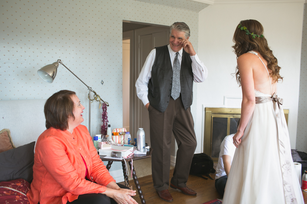 A bride in a white dress stands smiling with a man in a suit and vest and a woman in an orange blouse sitting on a bed, all sharing a light moment in a cozy room with a fireplace. Jessica Hill Photography - Wedding Photographer - Oregon