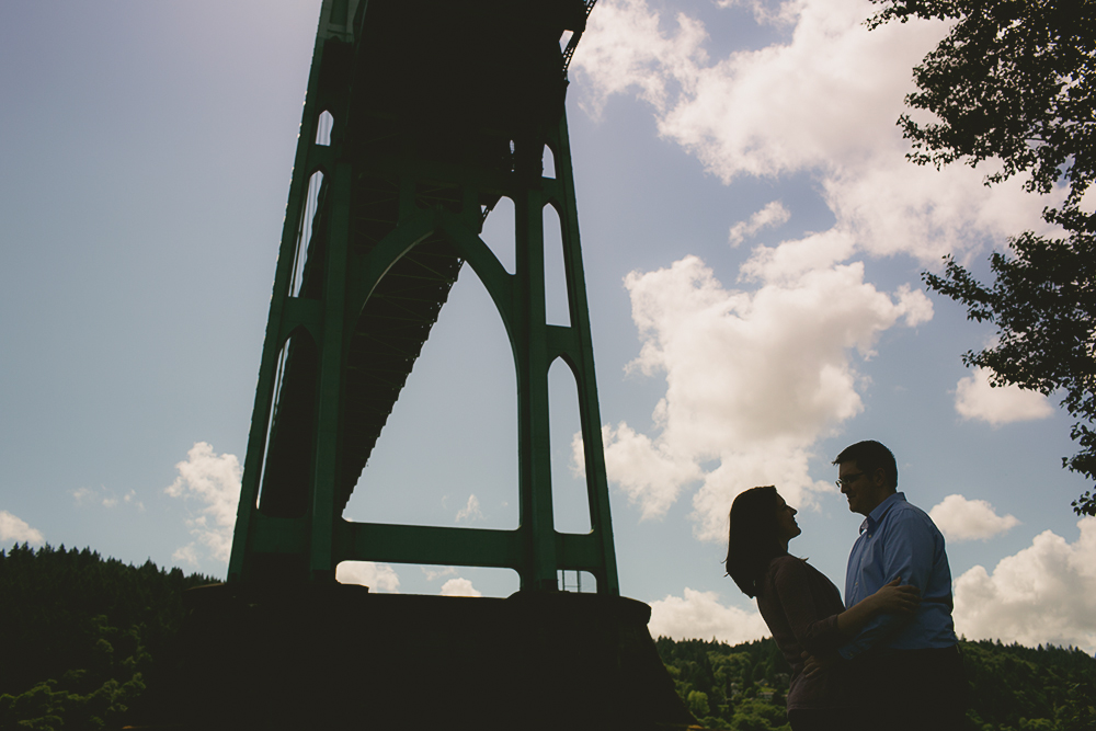 A couple stands under a large bridge, silhouetted against a partly cloudy sky, with trees and greenery in the background. Jessica Hill Photography - Wedding Photographer - Oregon