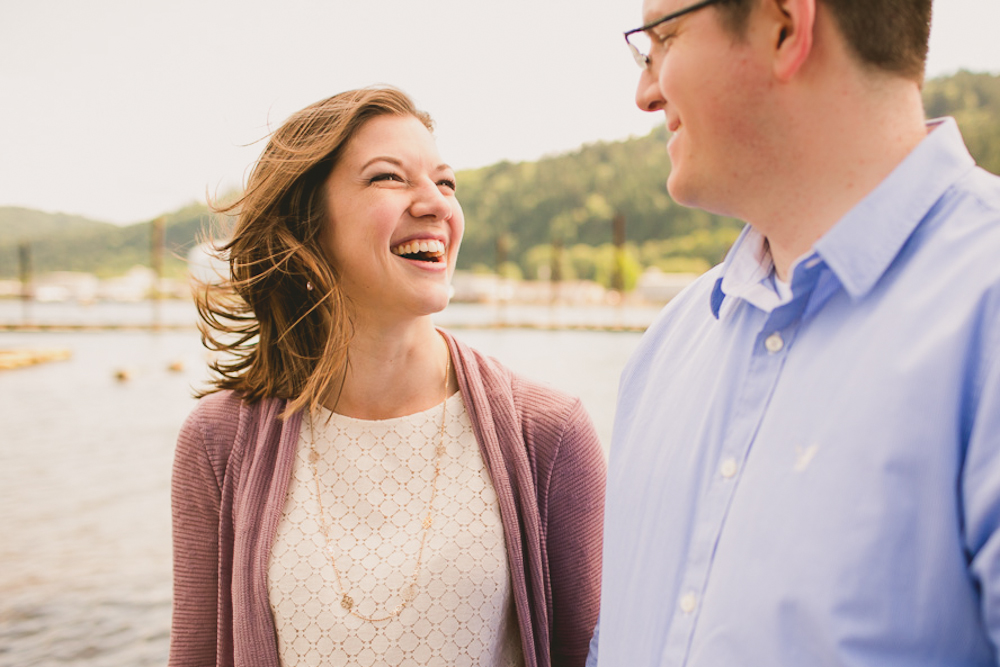 A woman laughing while standing next to a man in a light blue shirt, outdoors by the water with hills in the background. Jessica Hill Photography - Wedding Photographer - Oregon