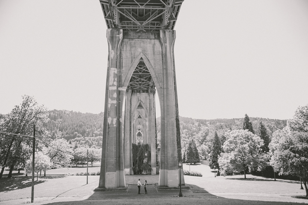 Two people stand under a large, arched concrete bridge in a park setting with trees and hills in the background. Jessica Hill Photography - Wedding Photographer - Oregon
