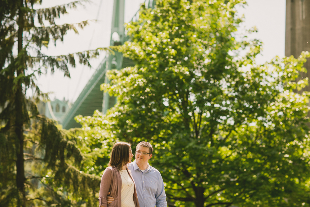 A couple stands together in a sunlit park with green trees and a large bridge structure in the background. Jessica Hill Photography - Wedding Photographer - Oregon