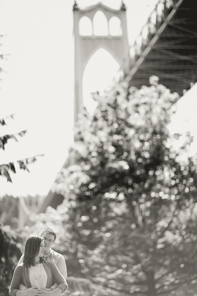 A couple stands close together outdoors beneath a large bridge, with trees and the bridge's structure prominent in the background. Jessica Hill Photography - Wedding Photographer - Oregon
