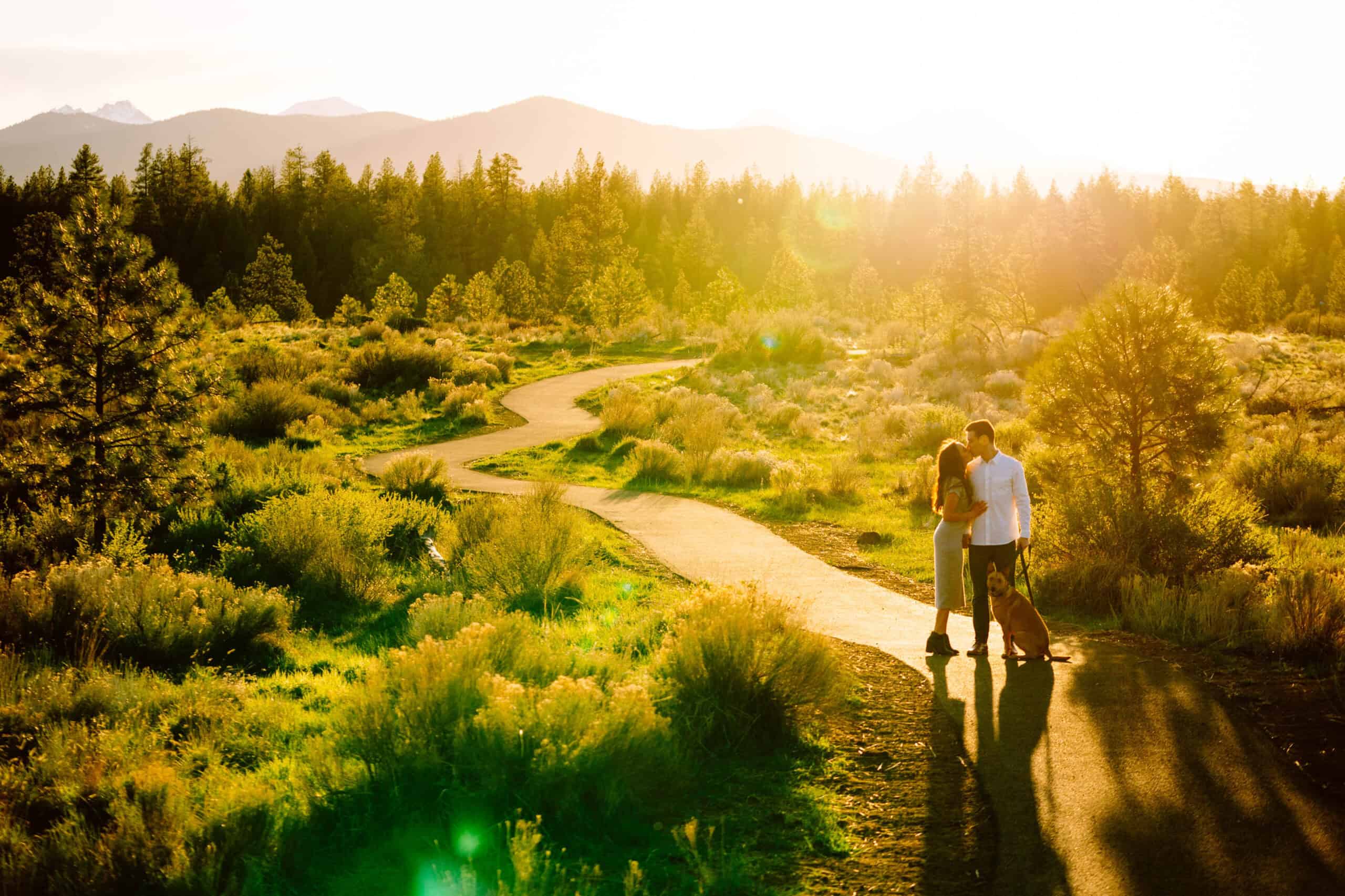 A couple stands on a winding paved path in a sunlit, grassy landscape with trees and mountains in the background. Jessica Hill Photography
