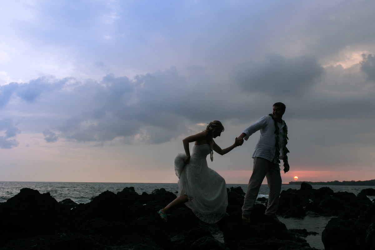 A bride and groom stand on rocky terrain by the ocean at sunset, with the groom helping the bride balance as they walk. The sky is cloudy with a hint of orange from the setting sun. Jessica Hill Photography - Wedding Photographer - Oregon