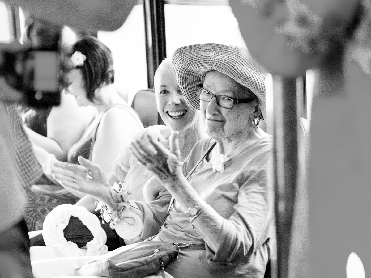 Two women sit closely together, smiling and clapping. One wears glasses and a wide-brimmed hat; the other has a corsage on her wrist. They appear happy and engaged, surrounded by other people in a bright setting. Jessica Hill Photography - Wedding Photographer - Oregon