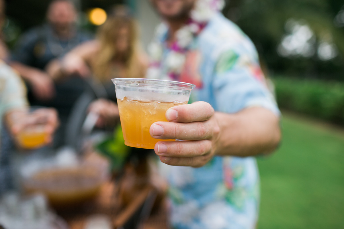 A person in a colorful, floral shirt holds a plastic cup filled with an orange drink toward the camera at an outdoor gathering, with people and greenery blurred in the background. Jessica Hill Photography - Wedding Photographer - Oregon