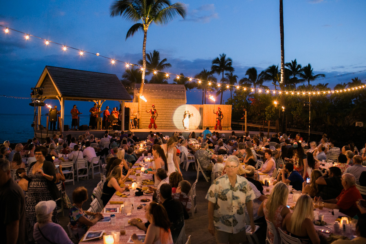 An outdoor luau at dusk shows a large crowd seated at tables with string lights overhead, watching performers on a stage near palm trees and the ocean. Jessica Hill Photography - Wedding Photographer - Oregon