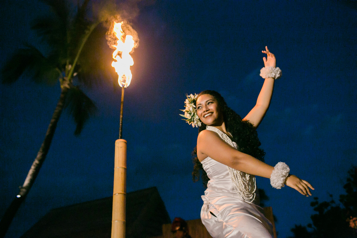 A woman in a white dress with floral accessories dances joyfully at night near a tall bamboo torch with a flame, set against a dark blue sky and palm trees. Jessica Hill Photography - Wedding Photographer - Oregon