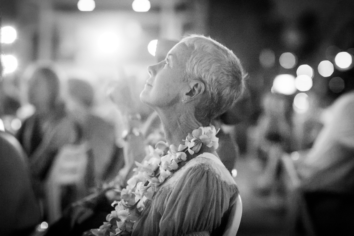 A woman with short gray hair wearing a flower lei looks upward thoughtfully in a dimly lit, blurred, indoor setting with other people in the background. The image is in black and white. Jessica Hill Photography - Wedding Photographer - Oregon