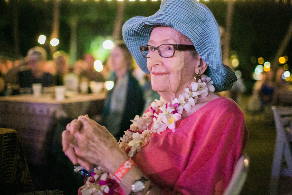 An elderly woman wearing a blue sunhat, glasses, a pink top, and a flower lei sits outdoors at night, smiling with her hands clasped. String lights and people are in the softly blurred background. Jessica Hill Photography - Wedding Photographer - Oregon