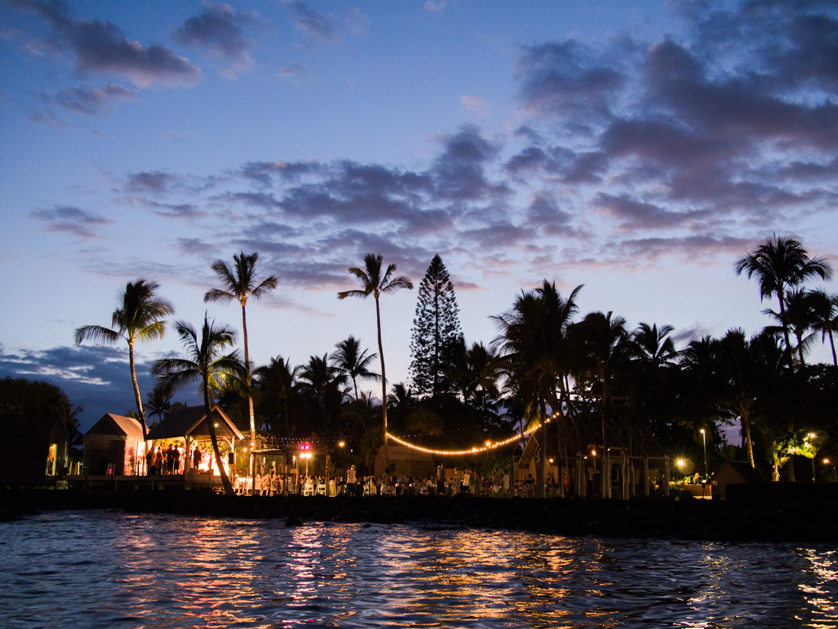 A tropical outdoor event at dusk, with palm trees silhouetted against a colorful sky and string lights illuminating people gathered near the waterfront. The water reflects the lights and the vibrant evening sky. Jessica Hill Photography - Wedding Photographer - Oregon