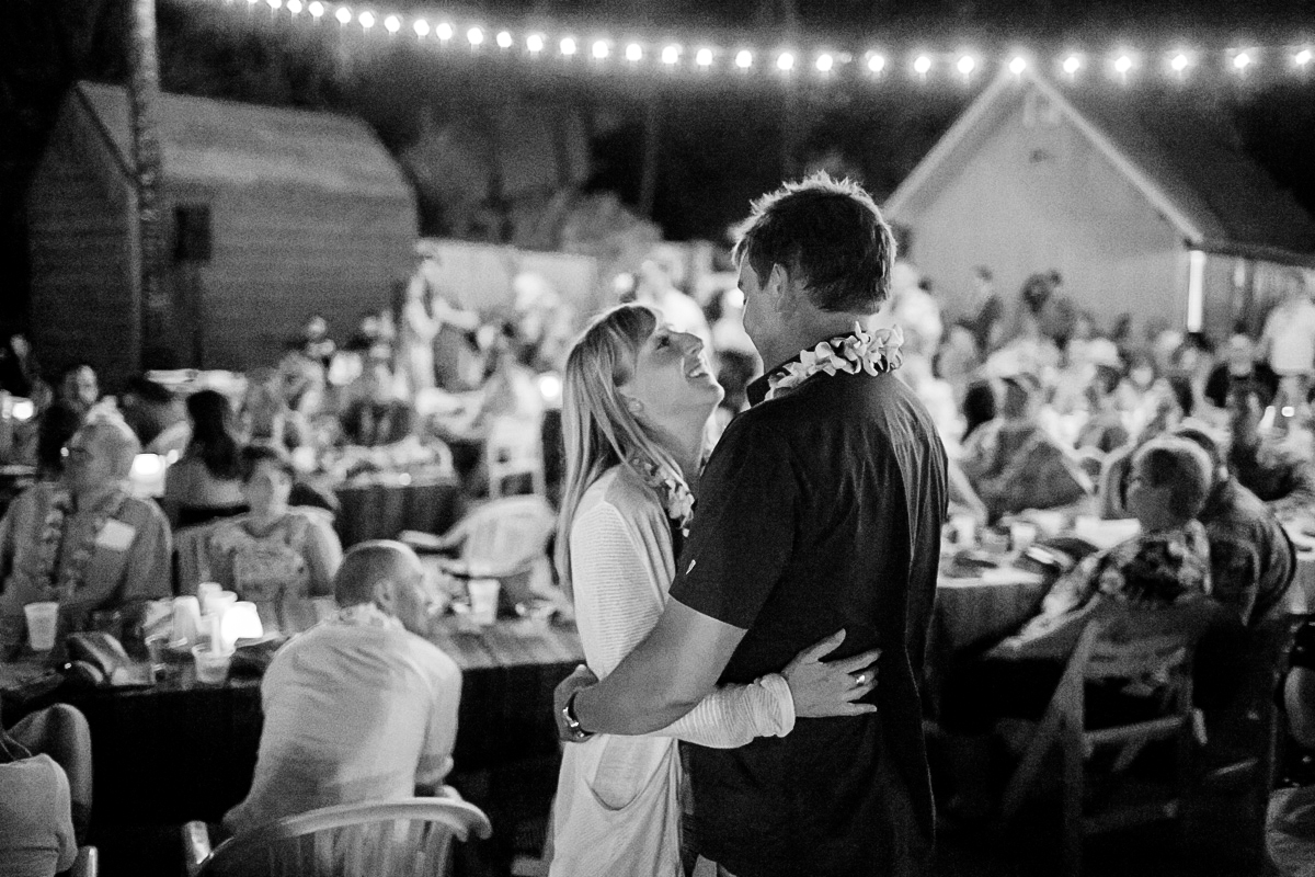 A couple smiles and embraces under string lights at an outdoor nighttime gathering, surrounded by seated guests at tables in a lively, festive atmosphere. Jessica Hill Photography - Wedding Photographer - Oregon