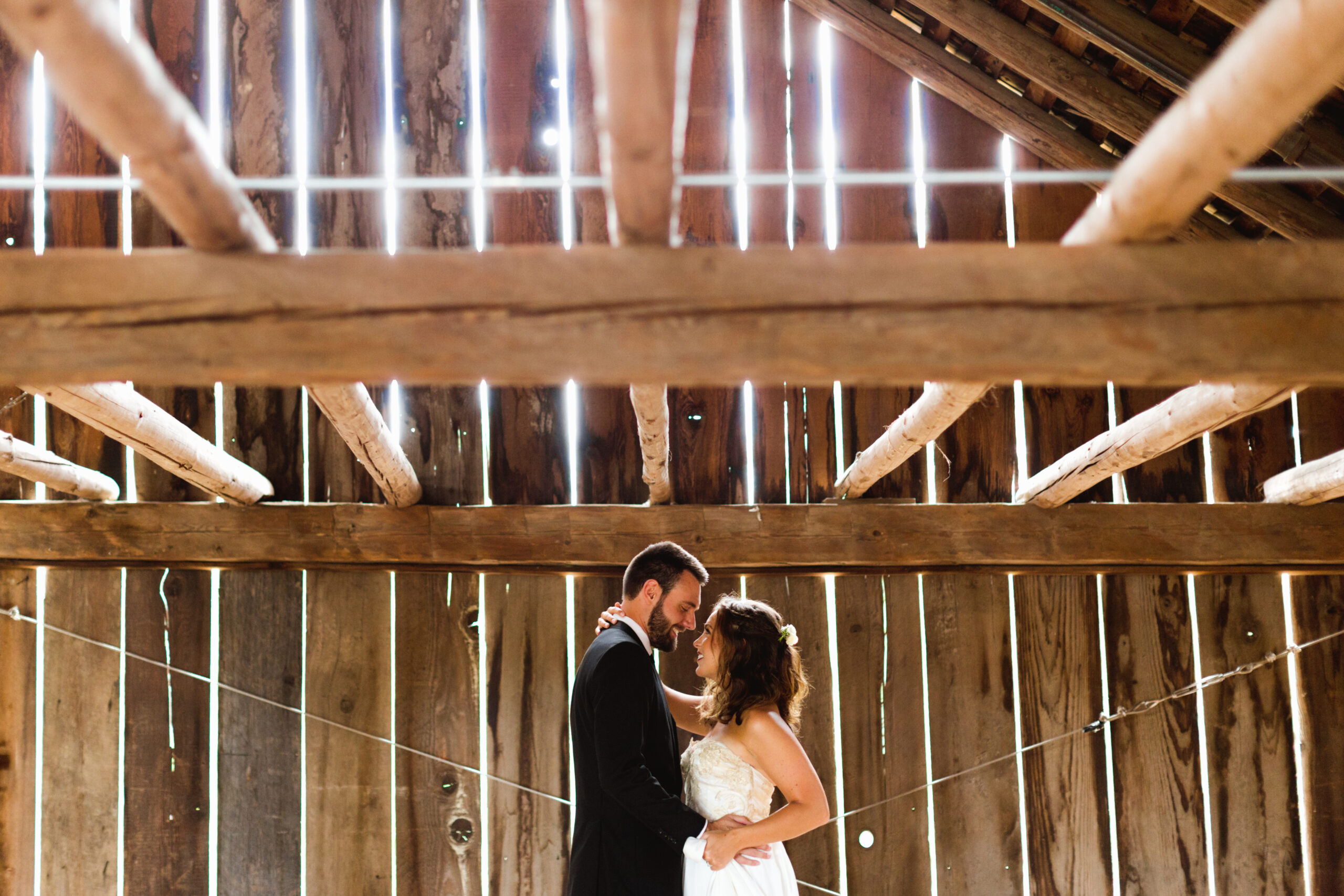 A bride and groom stand close together, embracing and gazing at each other in a rustic wooden barn with sunlight streaming through the gaps in the walls and roof beams above them. Jessica Hill Photography - Wedding Photographer - Oregon