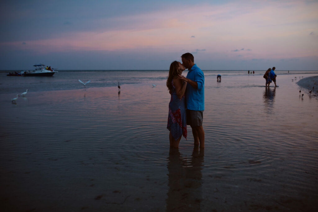 A couple stands close together in shallow water on a beach at sunset, with birds nearby and people and a boat in the background. Jessica Hill Photography