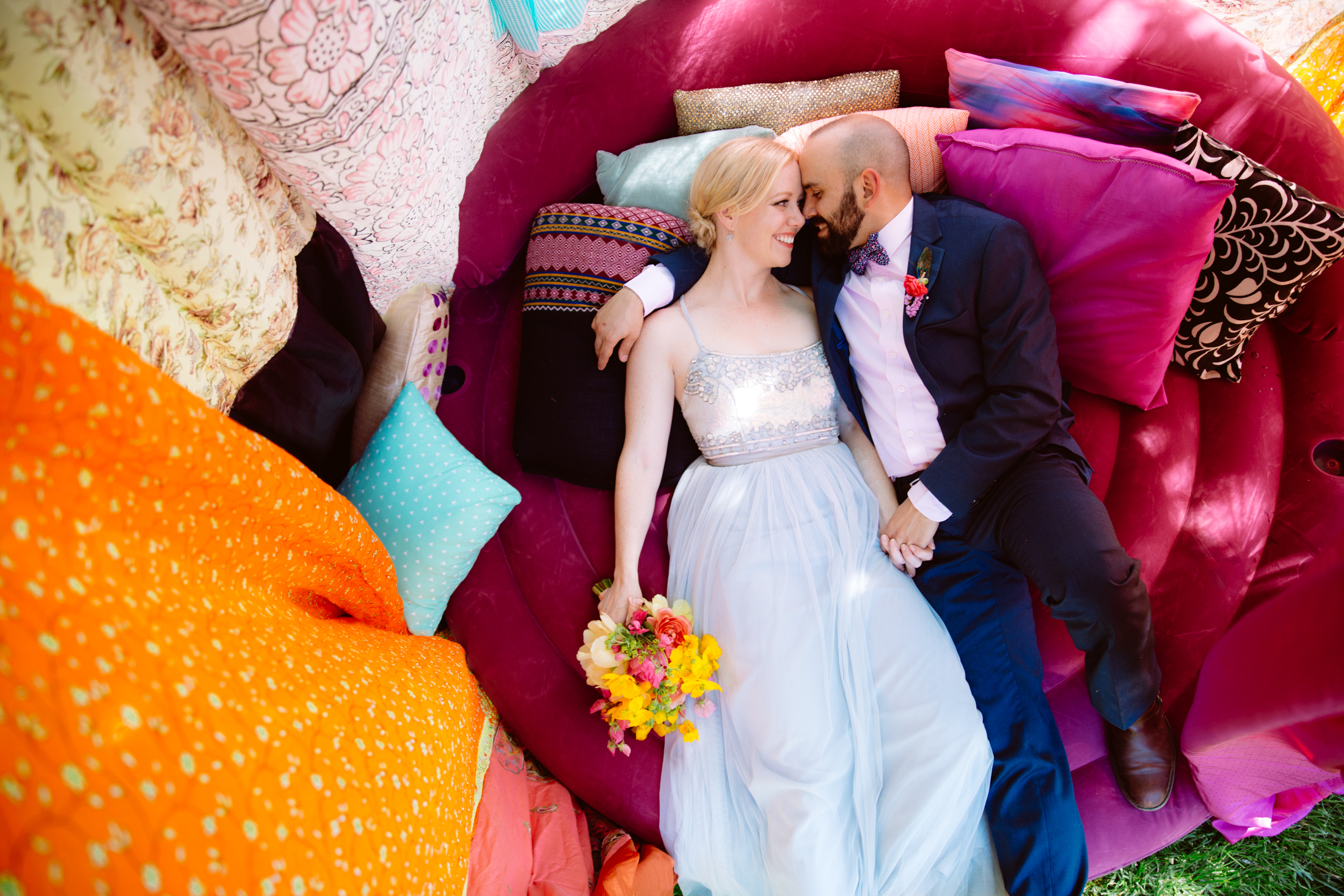 A couple in formal attire lies on colorful cushions outdoors, holding hands and smiling at each other; the woman holds a bouquet of flowers. Jessica Hill Photography