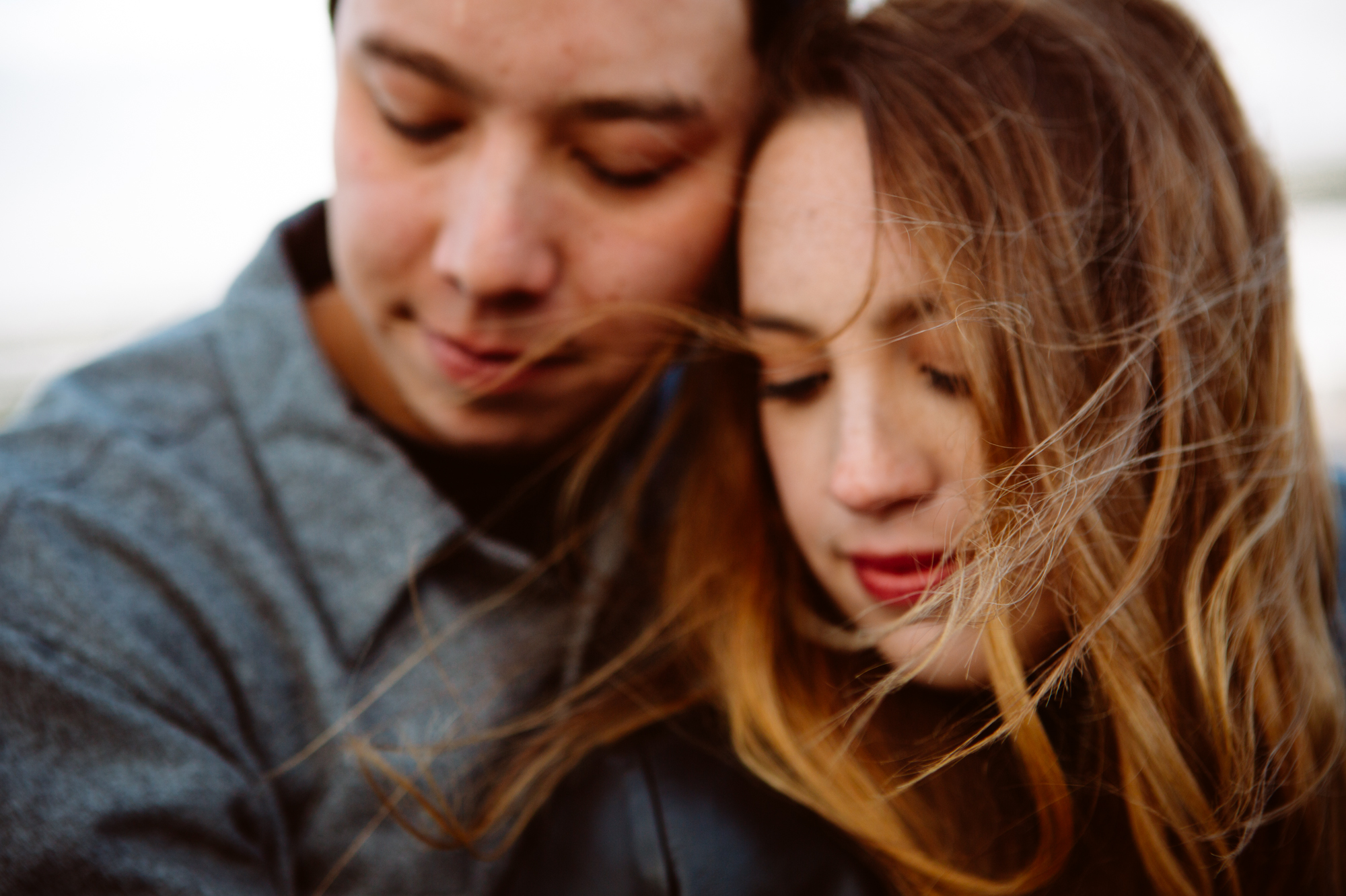 Close-up of two people with closed eyes leaning their heads together, with wind blowing the woman's hair across her face. Jessica Hill Photography