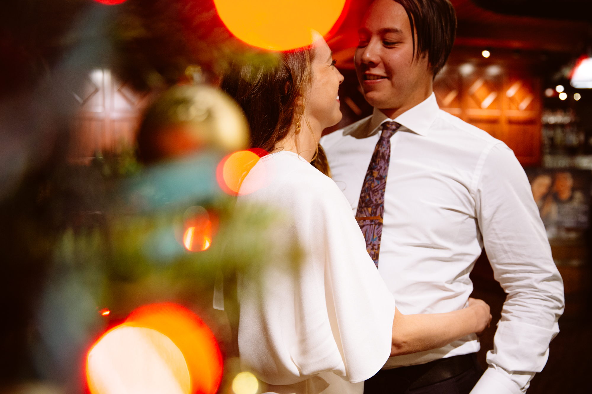 A couple dressed in formal attire stands close together, smiling at each other, with a decorated Christmas tree and blurred lights in the foreground. Jessica Hill Photography