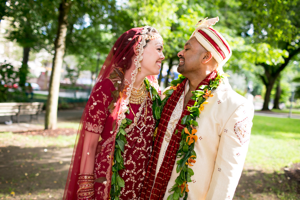A couple in traditional Indian wedding attire smile at each other outdoors, wearing garlands and standing close together, surrounded by green trees and sunlight. Jessica Hill Photography - Wedding Photographer - Oregon