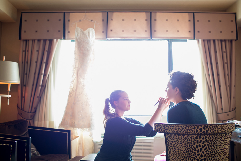 A makeup artist applies makeup to a seated woman in front of a bright window with a wedding dress hanging above. The scene is set in a warmly lit room with patterned curtains and a leopard-print chair. Jessica Hill Photography - Wedding Photographer - Oregon