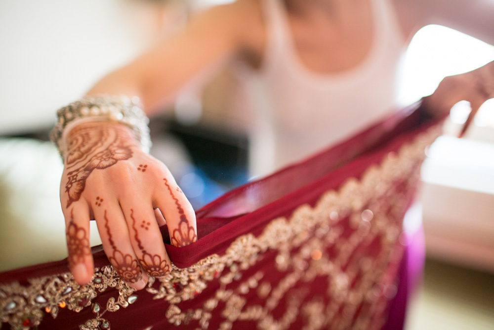 A person with intricate henna designs on their hand holds an ornate, red fabric decorated with gold embroidery and beadwork, preparing to drape it. The background is softly blurred. Jessica Hill Photography - Wedding Photographer - Oregon
