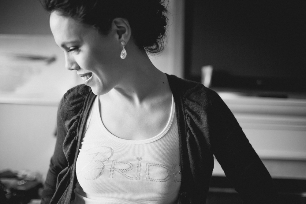 A woman smiles while looking down. She wears a tank top with "Bride" written on it and a dark cardigan, along with drop earrings. The photo is in black and white. Jessica Hill Photography - Wedding Photographer - Oregon