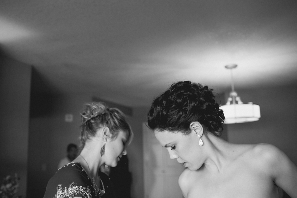 Two women in formal attire stand indoors, looking down. The woman in the foreground has short curly hair and earrings, while the woman behind her has lighter hair in an updo. The scene is softly lit with a ceiling light above. Jessica Hill Photography - Wedding Photographer - Oregon