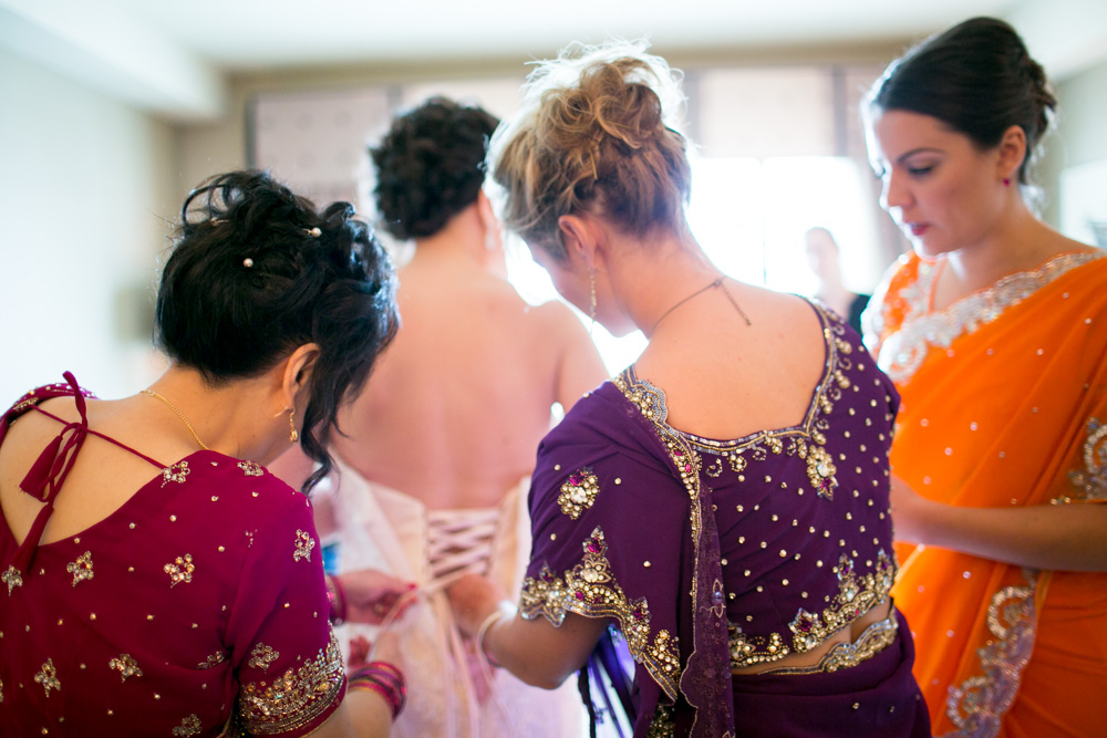 Three women in colorful sarees help another woman, who is wearing a white dress, lace up the back of her gown in a bright room, preparing for a special occasion. The scene is filled with anticipation and focus. Jessica Hill Photography - Wedding Photographer - Oregon