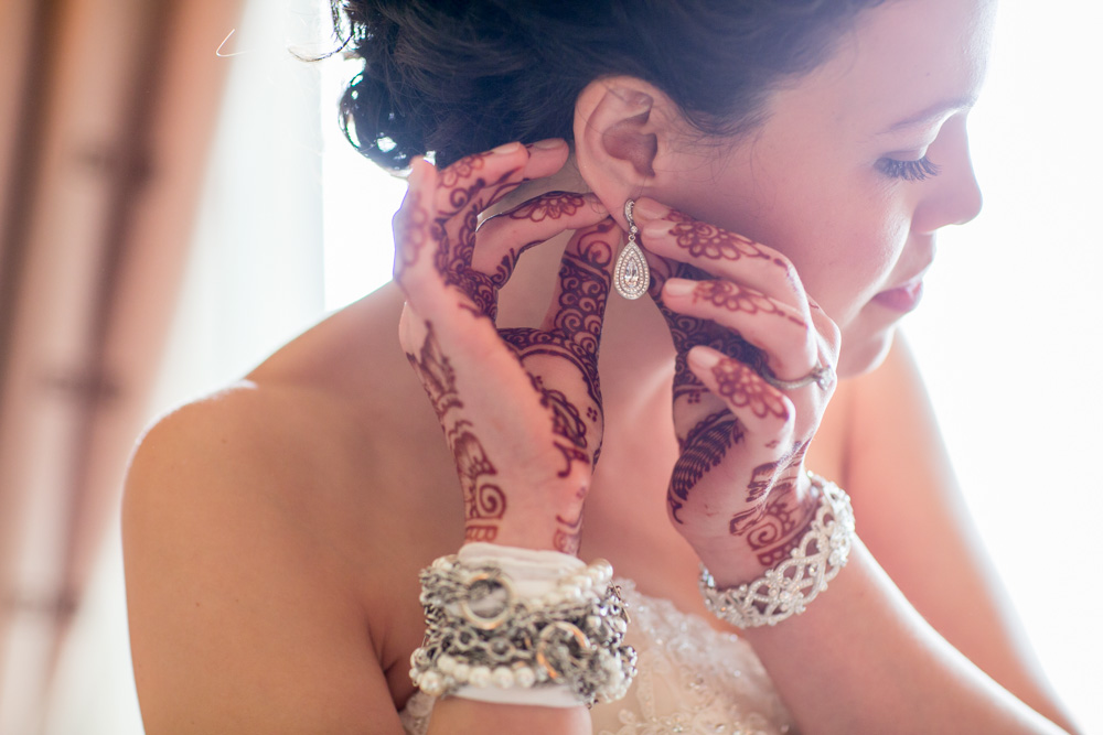 A bride with intricate henna designs on her hands and wrists adjusts a dangling earring, wearing multiple bracelets and a white dress, with soft natural light illuminating her face. Jessica Hill Photography - Wedding Photographer - Oregon