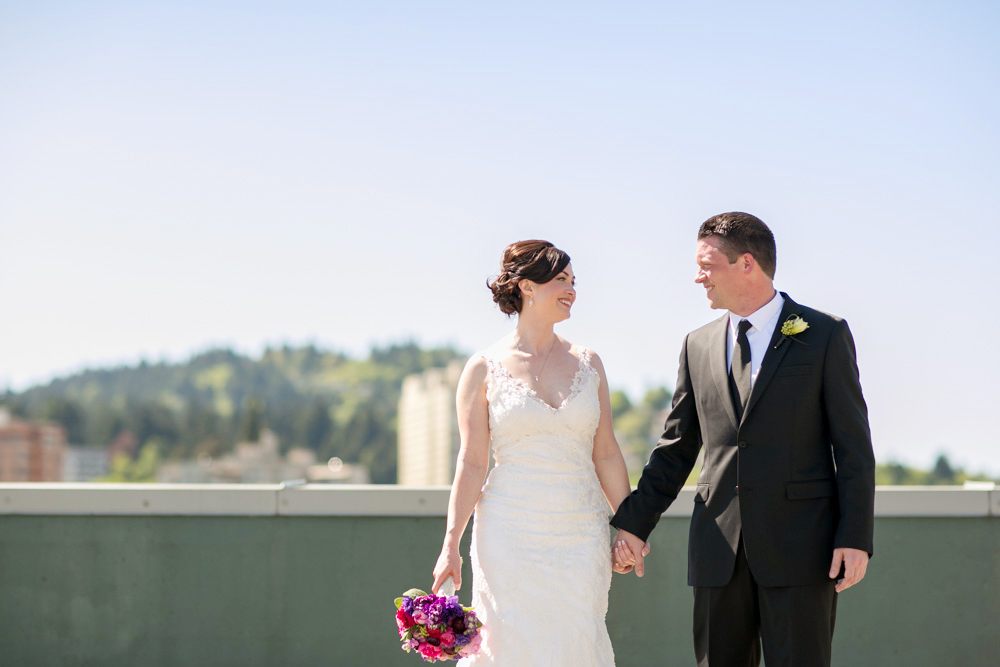 A bride in a white dress holding a bouquet and a groom in a black suit stand outdoors, holding hands and smiling at each other. Jessica Hill Photography - Wedding Photographer - Oregon