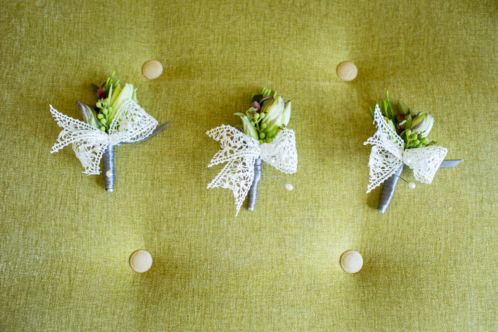 Three small boutonnieres with white lace bows are arranged in a row on a green, tufted fabric surface. Jessica Hill Photography - Wedding Photographer - Oregon