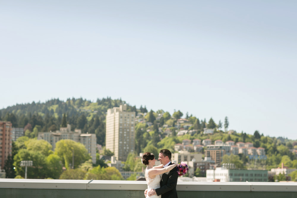A bride and groom stand on a rooftop embracing, with a cityscape and tree-covered hills in the background under a clear sky. Jessica Hill Photography - Wedding Photographer - Oregon