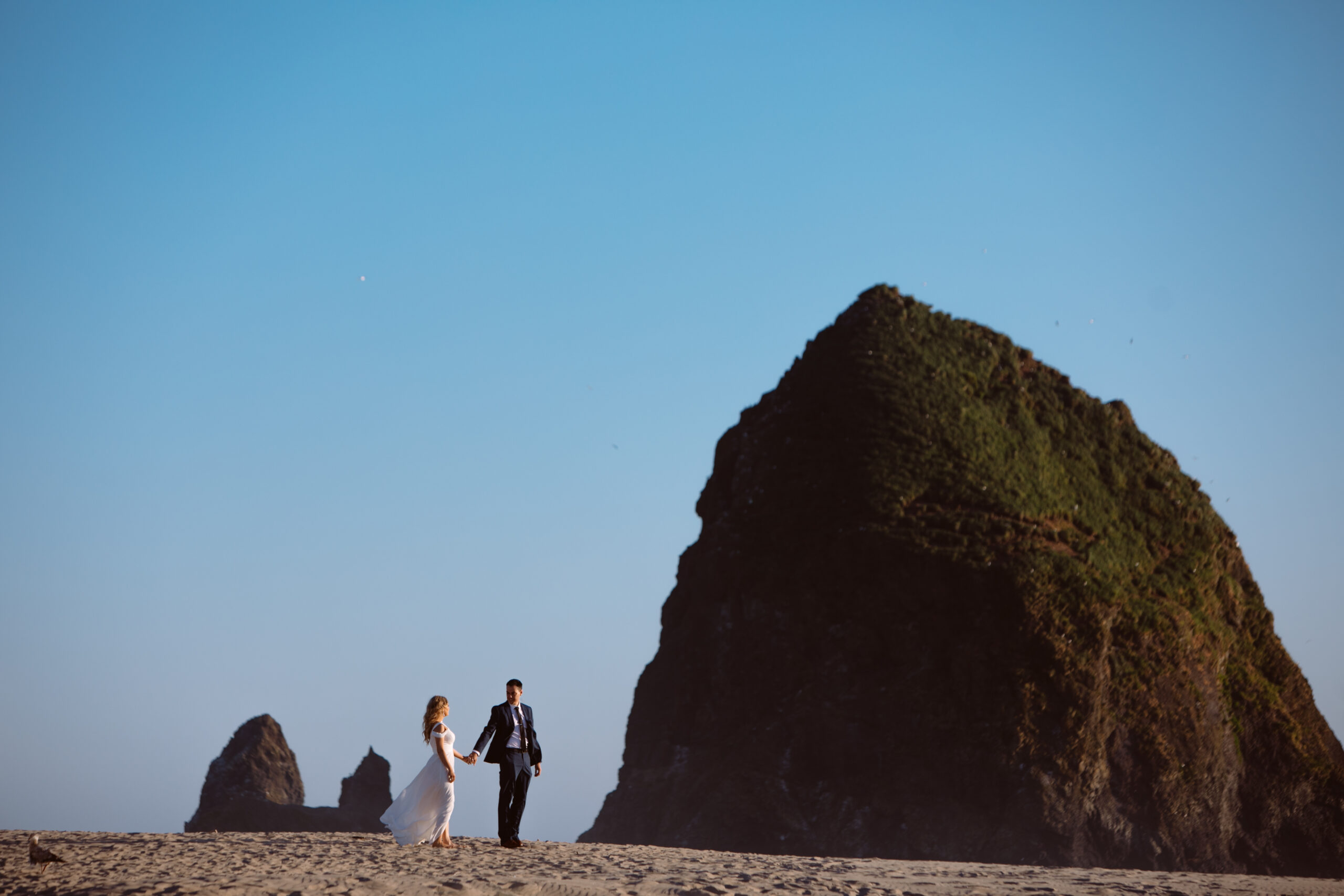 A couple in formal attire walks hand-in-hand on a sandy beach near large, grassy rock formations under a clear blue sky. Jessica Hill Photography