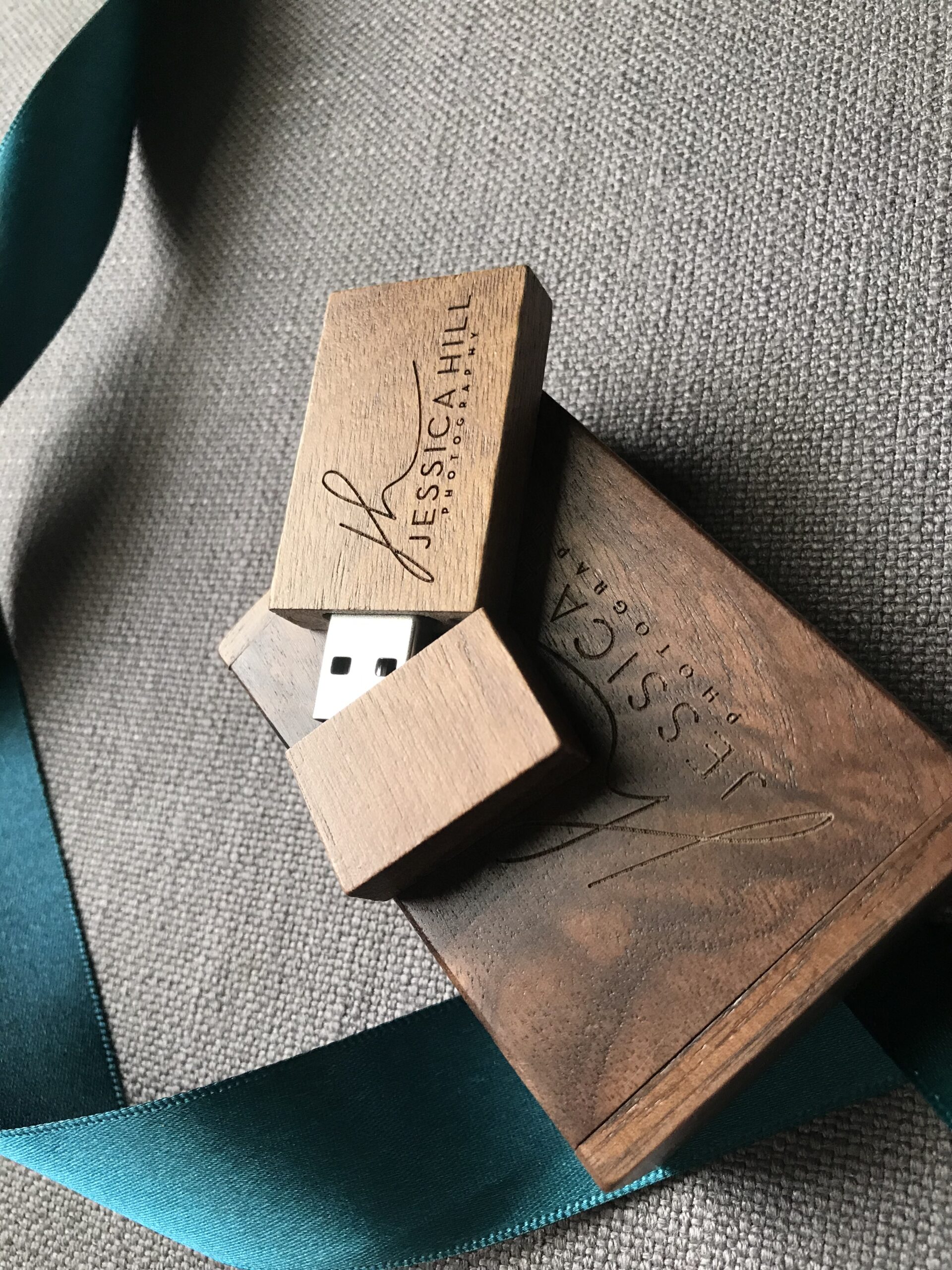 A wooden USB drive and box engraved with "Jessica Hill Photography" are shown on a gray fabric surface next to a teal ribbon. Jessica Hill Photography