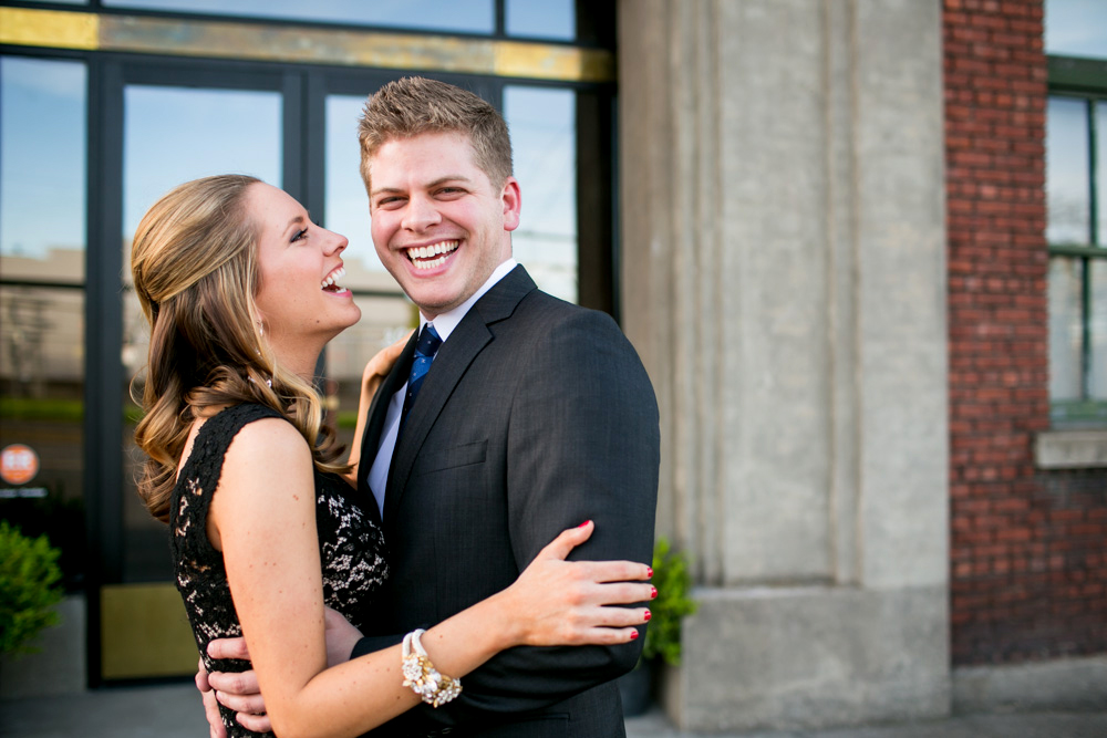 A woman in a black dress and a man in a suit smile and embrace each other outdoors in front of a building with large windows. Jessica Hill Photography - Wedding Photographer - Oregon