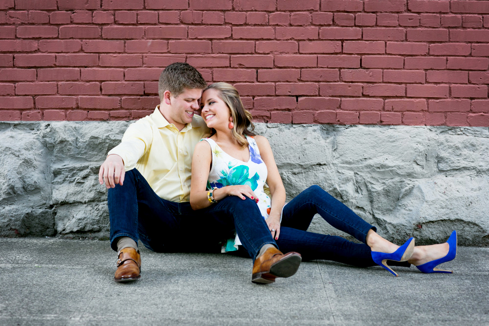 A man and woman sit on the sidewalk, leaning against a brick wall, smiling and leaning their heads together. Jessica Hill Photography - Wedding Photographer - Oregon