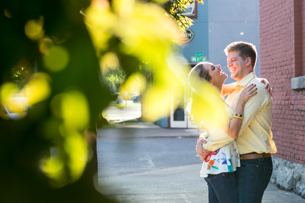 A couple stands on a sunlit sidewalk, smiling and embracing, with green leaves blurred in the foreground and a brick wall behind them. Jessica Hill Photography - Wedding Photographer - Oregon