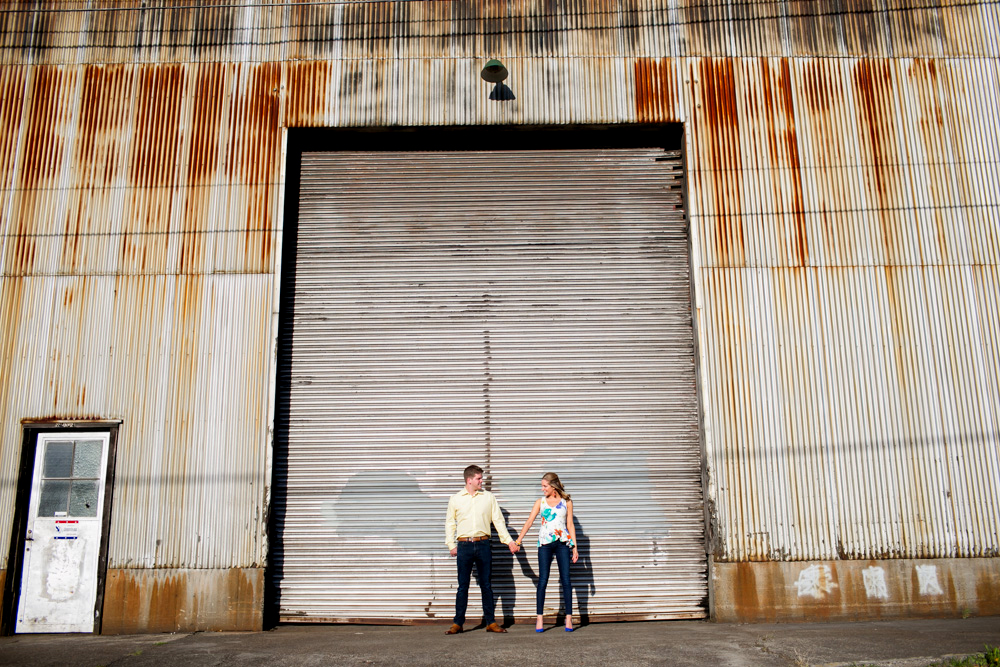 Two people stand holding hands in front of a large, corrugated metal industrial door with visible rust stains. Jessica Hill Photography - Wedding Photographer - Oregon