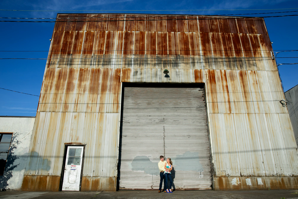 Two people stand close together in front of a large, rusted corrugated metal warehouse with a closed, roll-up door under a clear blue sky. Jessica Hill Photography - Wedding Photographer - Oregon