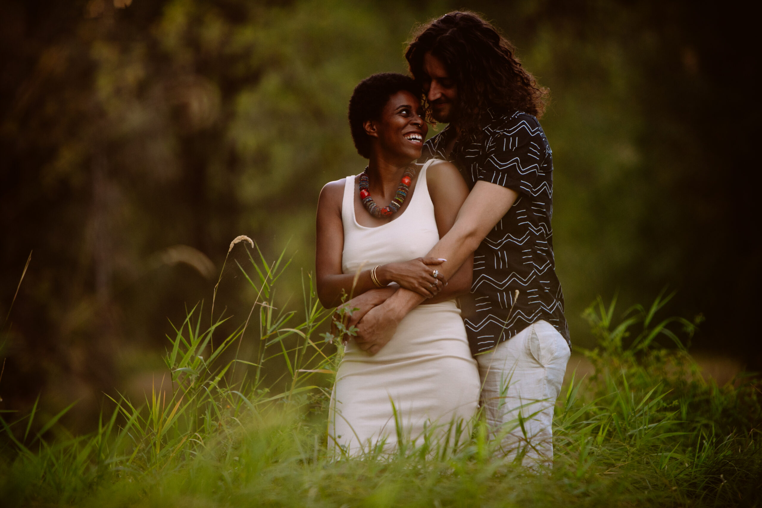 A couple stands together in a grassy field, smiling and embracing, with trees blurred in the background. Jessica Hill Photography