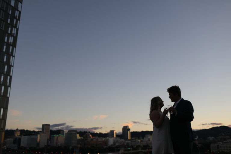A couple stands close together in formal attire, silhouetted against a city skyline at dusk with a clear sky above. Jessica Hill Photography