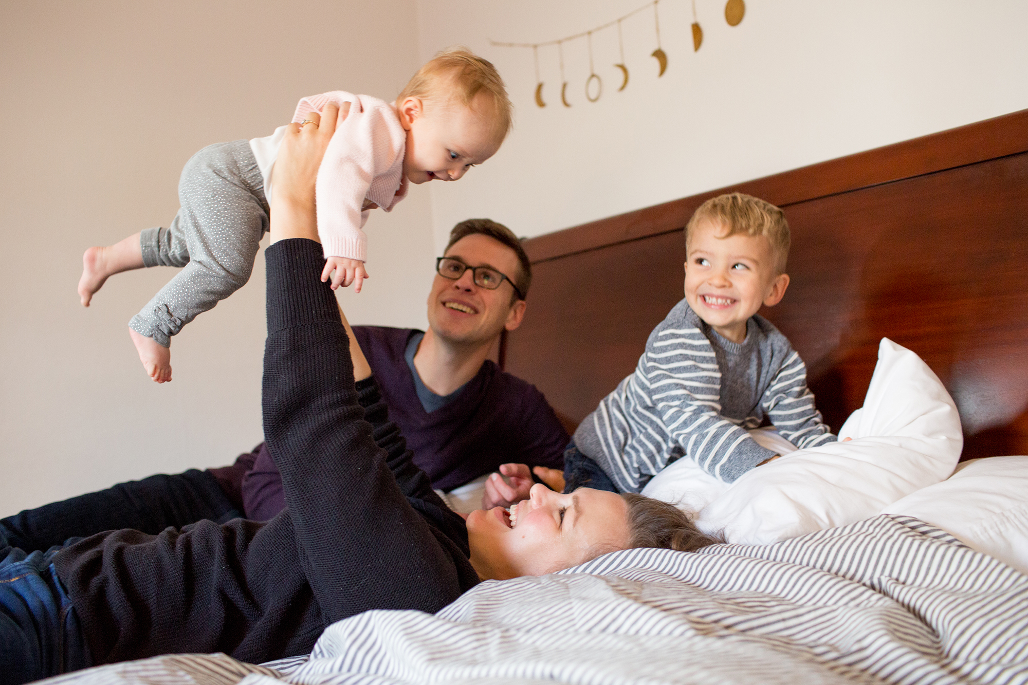Two adults and two young children play together on a bed; one adult lifts a baby while the other adult and a smiling child watch. Jessica Hill Photography