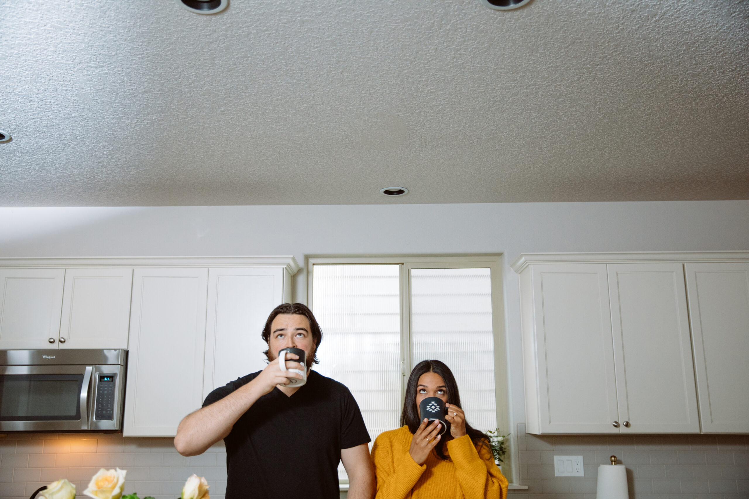 A man and woman stand in a kitchen, drinking from mugs. White cabinets and a microwave are visible in the background. Jessica Hill Photography